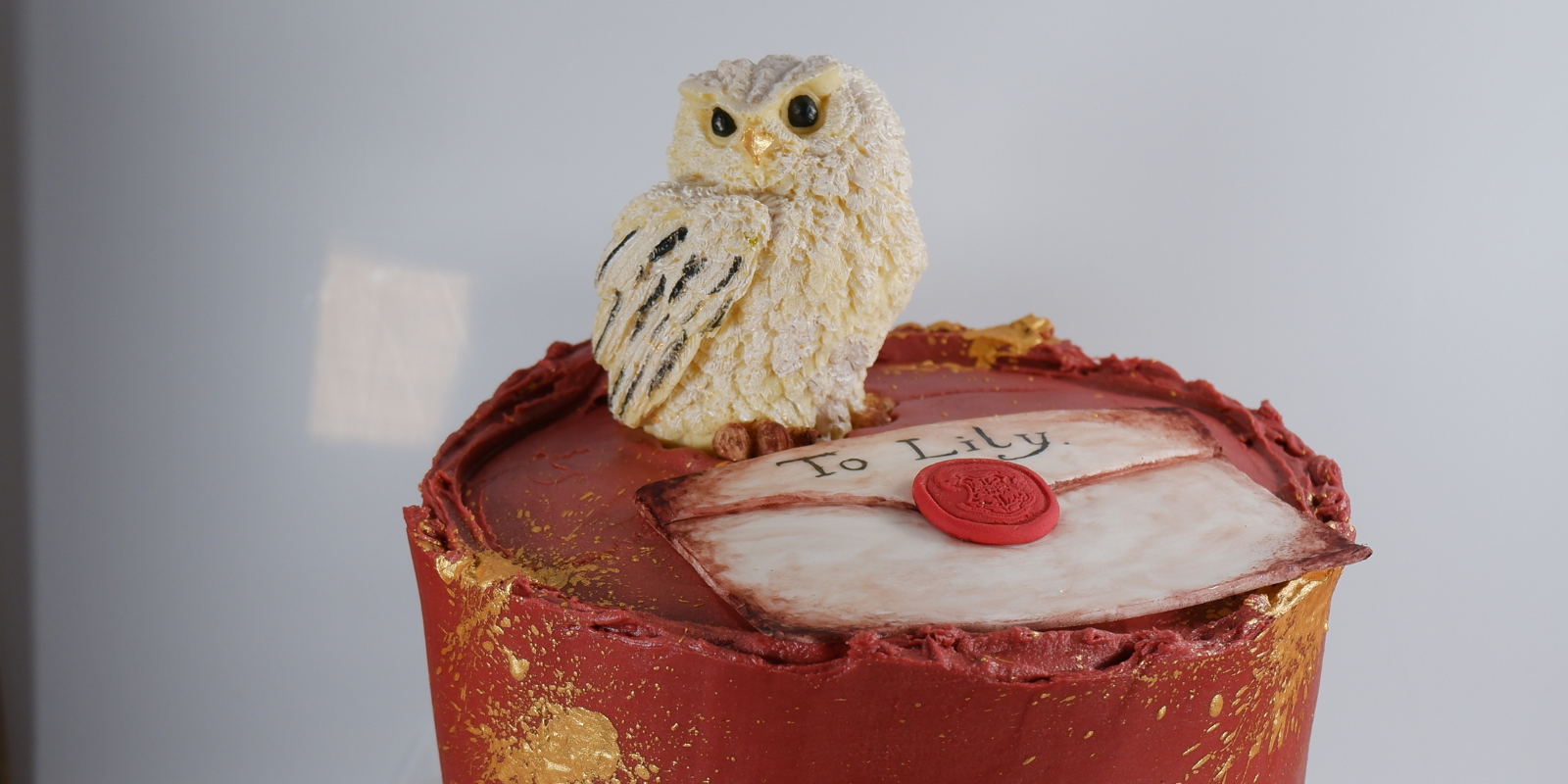 Cake with Harry Potter theme, Burgundy buttercream with gold splash, there is Hedwig on top with a old edible letter with the name of the child on the letter.