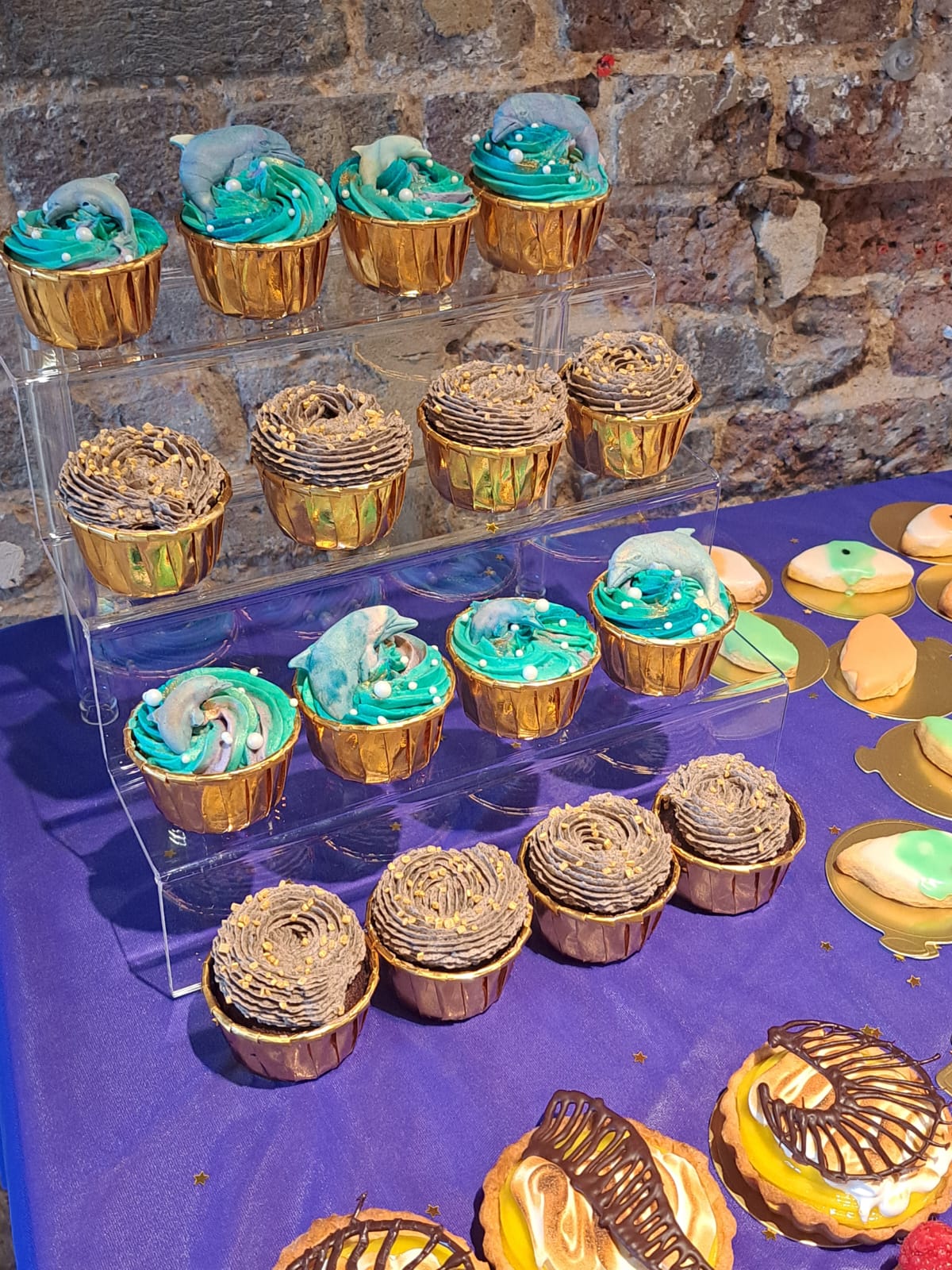 in the centre of the image are cupcakes, one with oreo buttercream and gold sprinkles and the other with blue buttercream and a chocolate dolphin on top to represent the art.